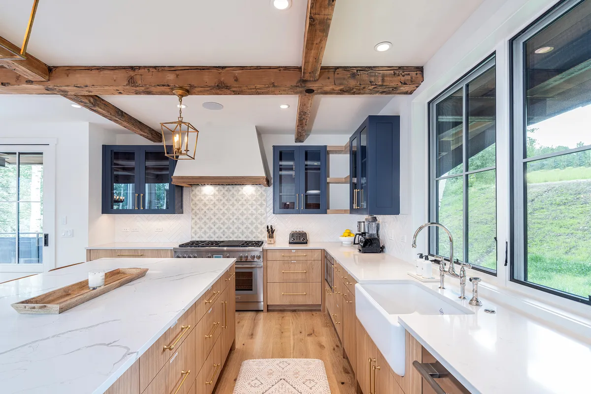 A bright, modern kitchen with a large kitchen island, stainless steel appliances, blue cabinetry, exposed wood beams, and expansive windows that let in natural light. The setup encourages cooking, entertaining, and sharing meals with a view.