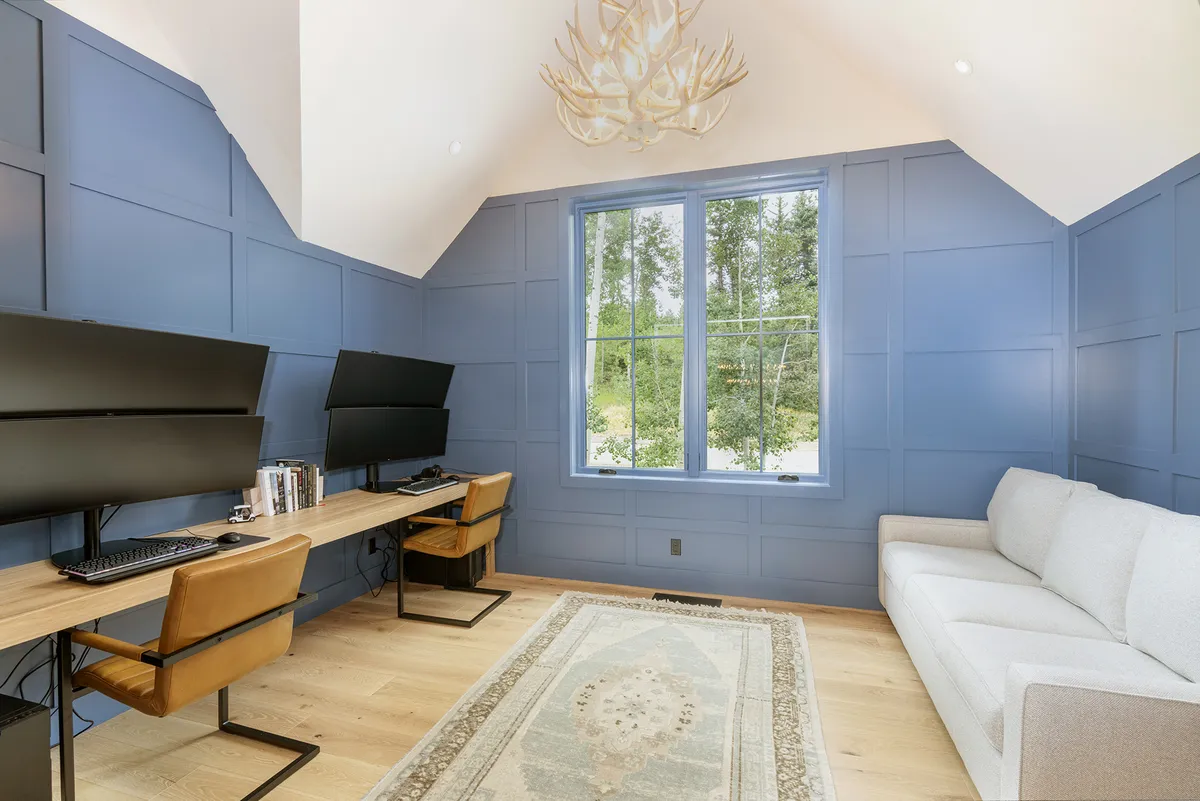 A stylish, expansive office/loft with blue paneled walls, large windows, modern desks equipped with monitors, an antler chandelier, and a light-colored comfy sofa. The room exudes productivity and calm, ideal for blending work and relaxation.