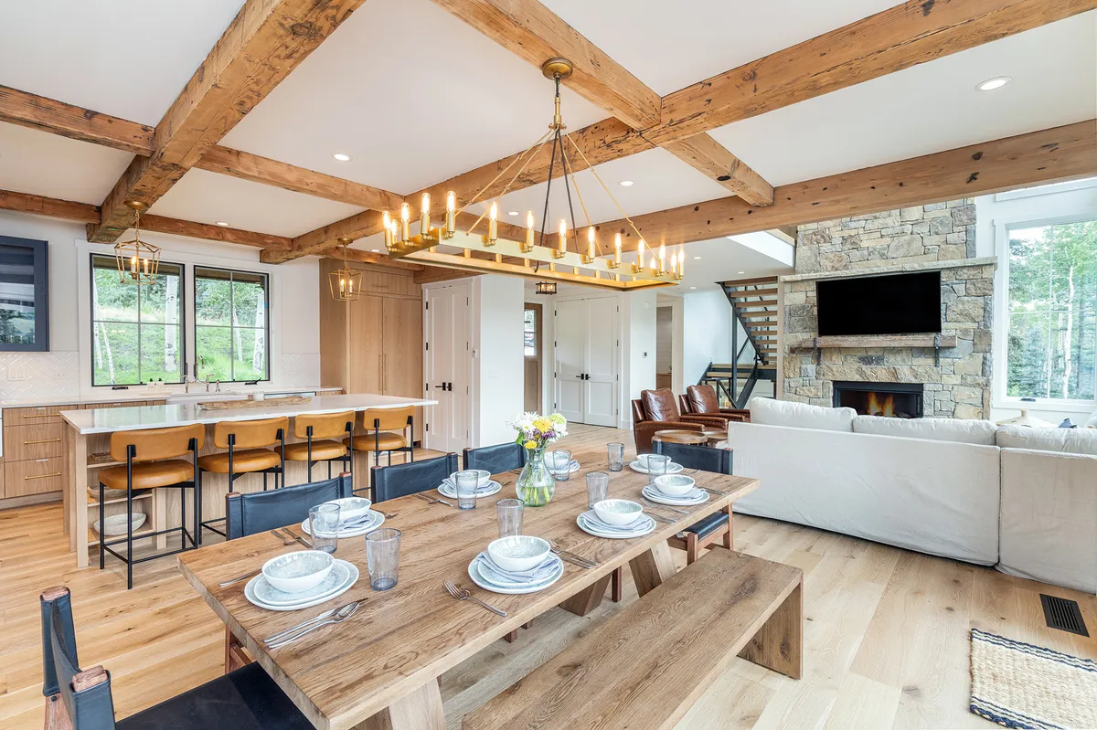 A warm and welcoming view of the dining area, with a standout rustic dining table, elegant chandelier, a kitchen island in the background, and a striking stone fireplace. The space is clearly designed for social gatherings, shared meals, and cozy evenings.