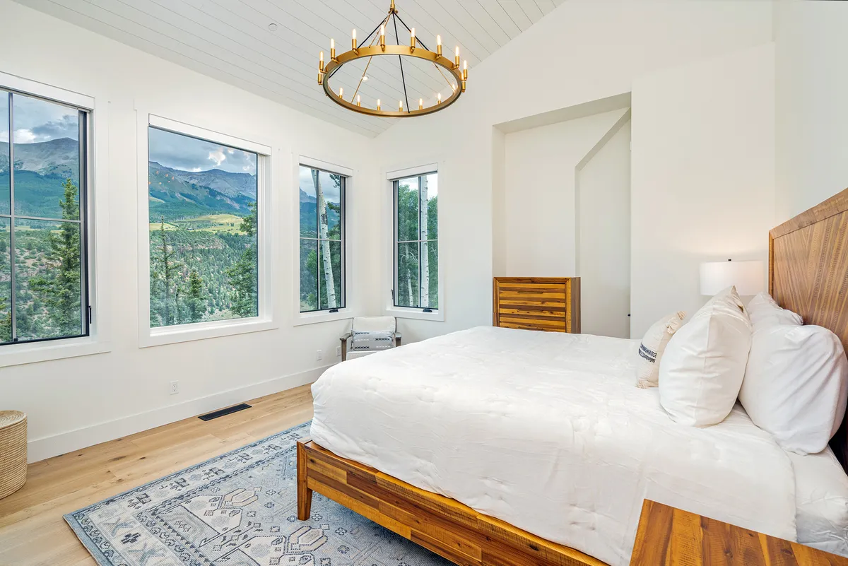This image showcases a king bedroom with a large bed adorned with decorative pillows, wooden furniture, and expansive windows that frame breathtaking mountain views. Natural light pours in, creating a serene and refreshing ambiance—ideal for rest.