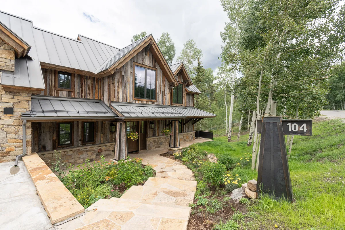 This image features the stone facade of the house, large windows, a welcoming pathway leading to the entrance, and a house number sign. The surrounding landscape includes trees and greenery, creating a serene, well-maintained first impression.