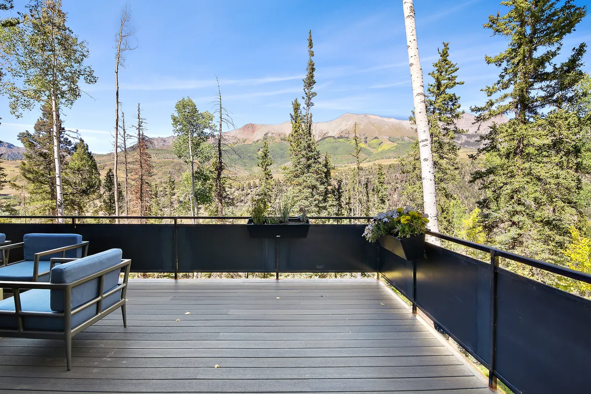 The main deck area offers outdoor seating surrounded by trees and framed by spectacular mountain views. Planters with flowers add a touch of color, and the space invites relaxation, conversation, and soaking in the natural beauty just outside the main living spaces.