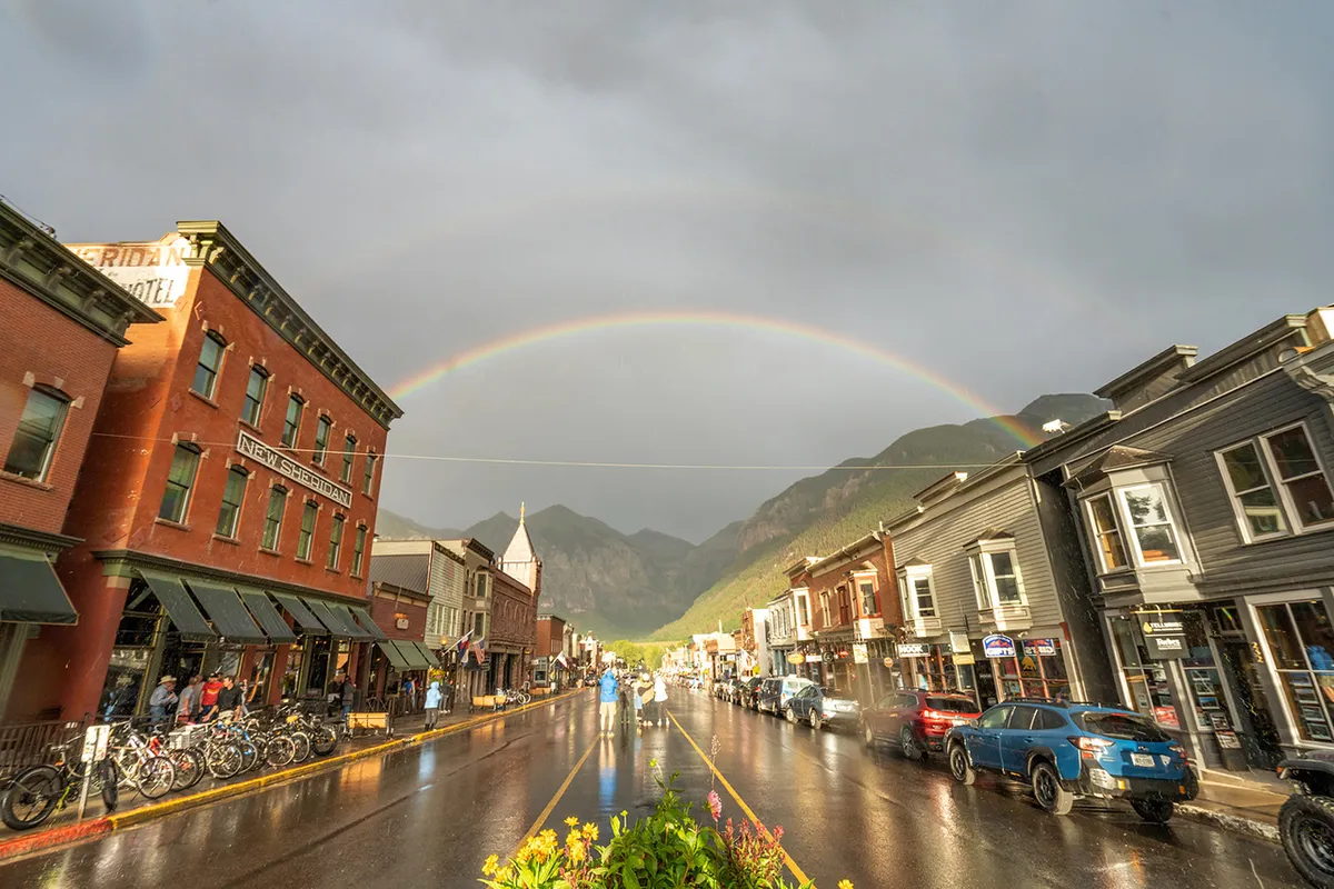 A vibrant town scene featuring historic buildings, a rainbow arching over wet streets, and families strolling or biking. The image captures the essence of exploration, shopping, and discovering local charm in Telluride.