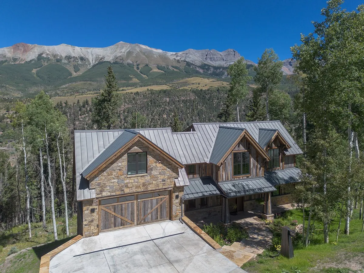 An expansive exterior view of the home's stone and wood facade, modern metal roof, wide driveway, and two-car garage, framed by surrounding trees and mountain views.