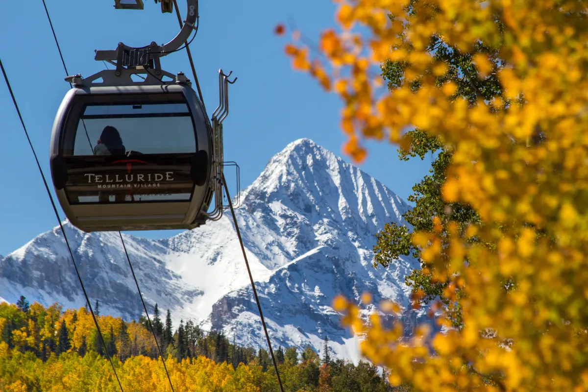 A panoramic view featuring a gondola gliding above a vibrant autumn landscape, snow-capped mountains in the background, and golden fall foliage below. The scene promises adventure and easy access to the outdoors, capturing the thrill of mountain exploration.