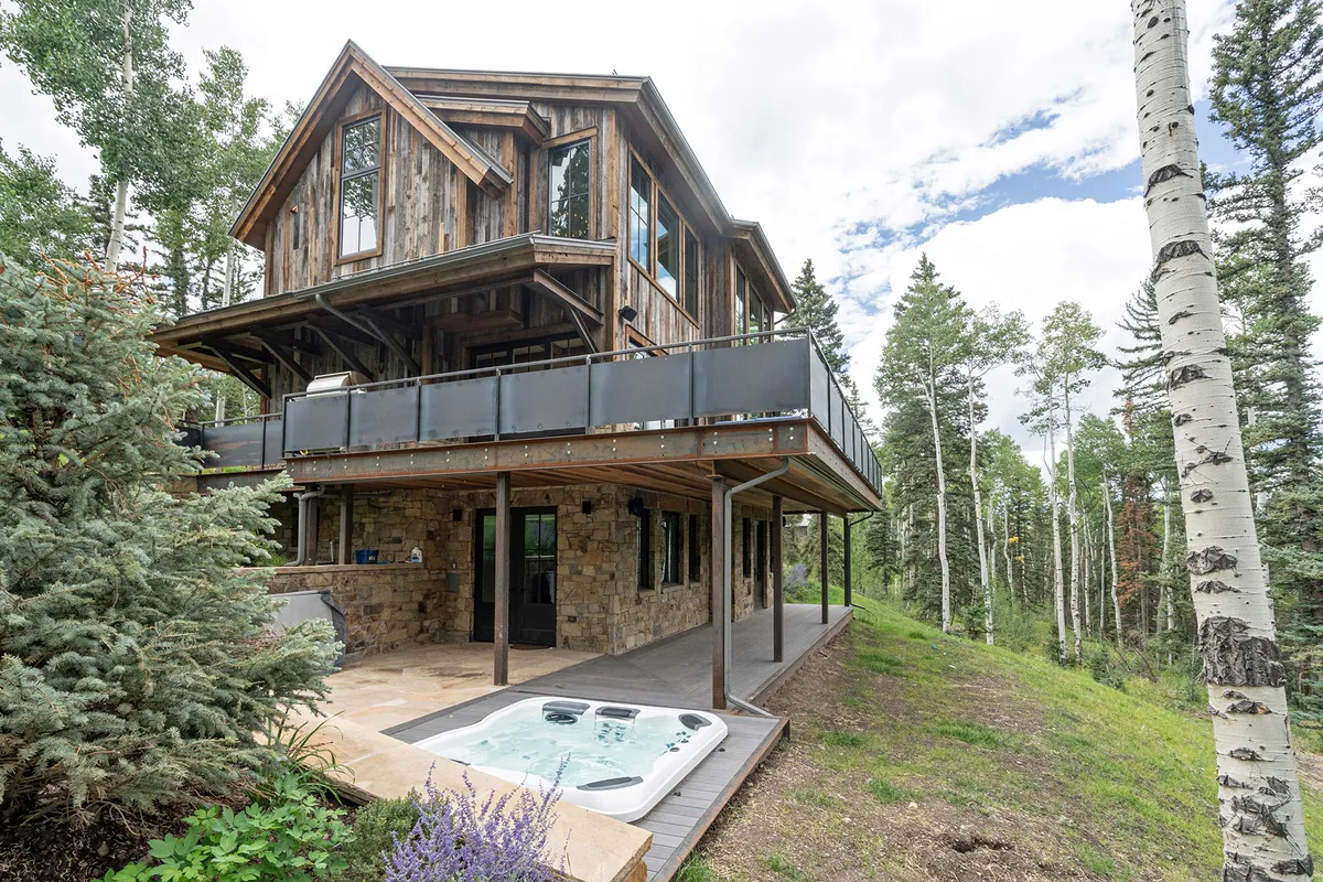 A spa sits on a wooden balcony flanked by stone details, with surrounding trees providing privacy and a view to nature. The mood is tranquil, highlighting both relaxation and connection with the outdoors.