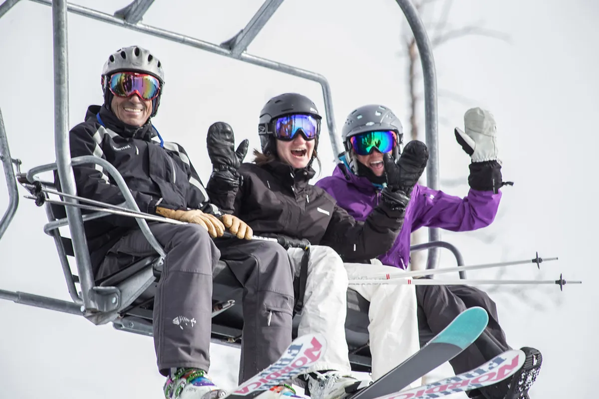 Three skiers are riding up a chairlift over a snow-covered mountain landscape, surrounded by evergreens and bright winter skies. Their colorful ski gear pops against the bright white snow, conveying the excitement of a day on the slopes.