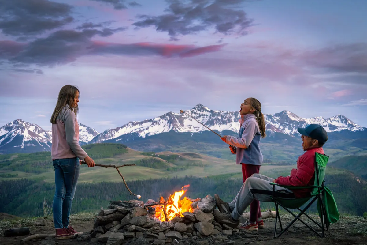 Children are gathered around a glowing campfire, roasting marshmallows against a stunning mountain backdrop. The atmosphere is lively and joyful, highlighting families spending quality time outdoors in a picturesque natural setting.