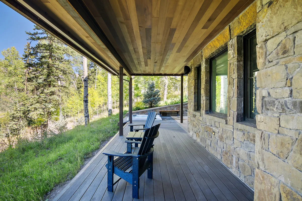 A peaceful wooden deck with rocking chairs sits beside a stone wall and is surrounded by leafy trees. The scene suggests moments of relaxation, reading, or socializing while immersed in nature.