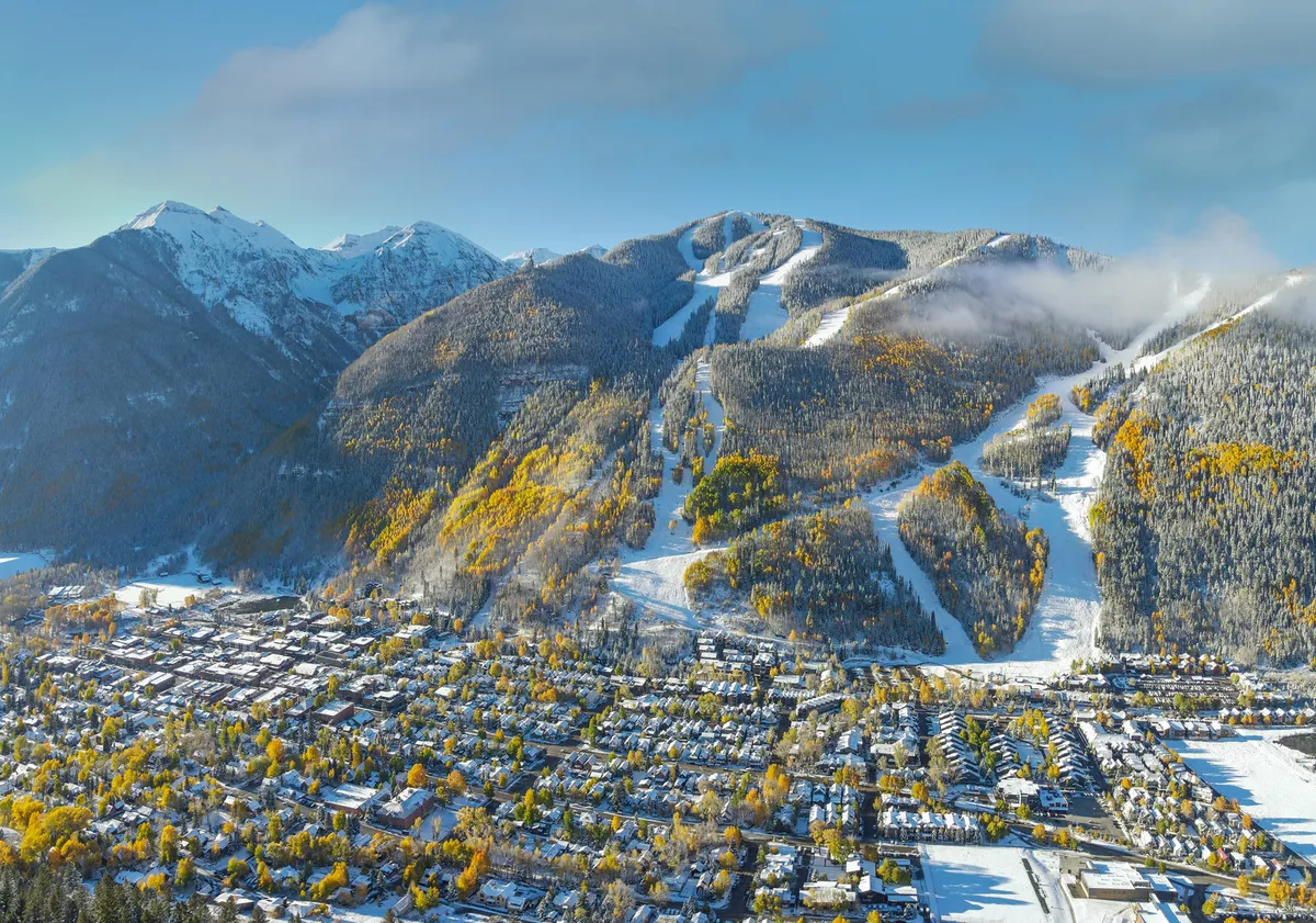 An expansive view of snow-capped mountains, colorful autumn trees, ski slopes, and the inviting town nestled below. The scene is energetic yet peaceful, reflecting both winter and summer activities available in Mountain Village.