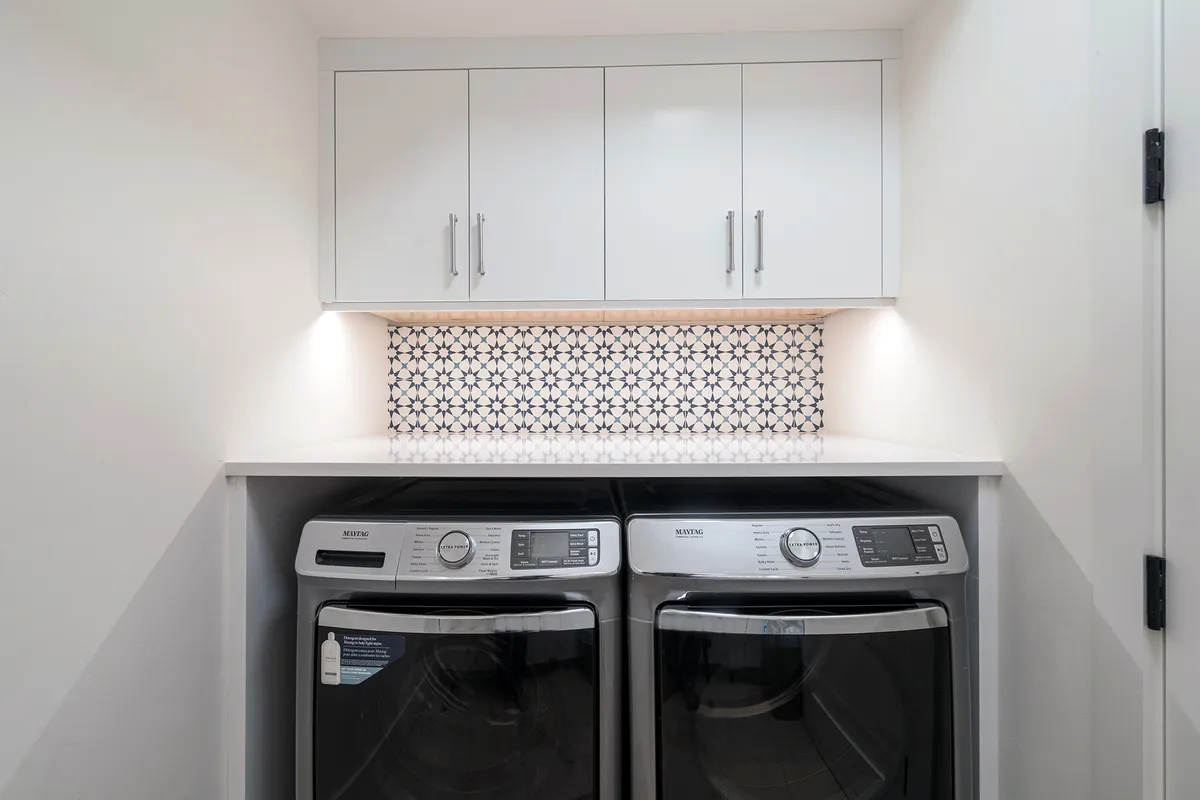 A modern laundry room with a washer and dryer set neatly into custom cabinetry, decorative backsplash, upper storage cabinets, and an organized, bright, and functional atmosphere.